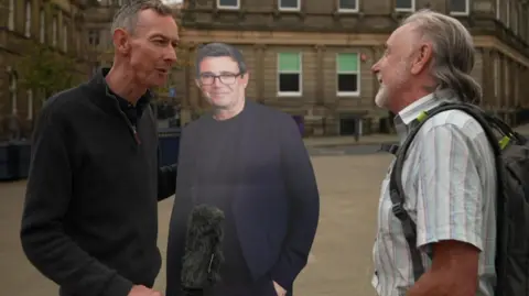 BBC Aman holding a microphone and cardboard cut out of Andy Burnham interviews a man in a white shirt on a street