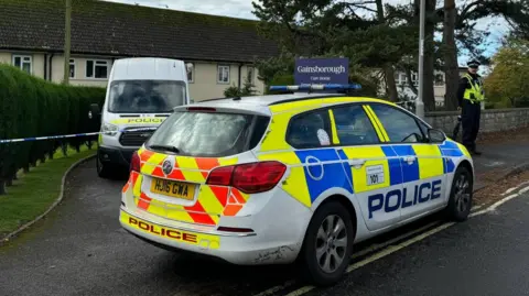 BBC A police car and van are parked either side of police tape at Gainsborough Care Home. A uniformed officer stands guarding the cordon.