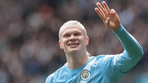 Erling Haaland, wearing Manchester City's light blue kit, celebrates his latest hat-trick by making a three-finger salute with his left hand