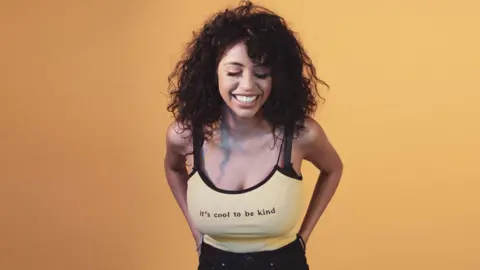 Ranvia Ranvia smiling in front of an orange background, where she is stood wearing a strappy vest top which says 'it's cool to be kind.' The photo was taken before her breast reduction surgery