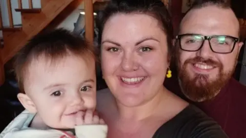 Family photo Rachel who had dark hair in a bun and rubber duck earrings in her ears, smiling and looking at the camera, with baby Eleanor in her arms and Chaz, who has glasses and a ginger beard, stood beside her.