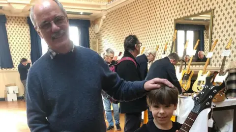 Family photo Lee stands with his hand on the head of a younger Olly who is holding an electric guitar.  In the background there are stalls selling guitars and people browsing. Lee is balding with white hair and glasses and wearing a round neck blue sweater, he is holding a guitar case. Olly is wearing a black long sleeve t-short, black jeans and grey trainers.