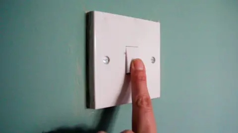 Close shot of a finger pressing a white light switch, against a green wall