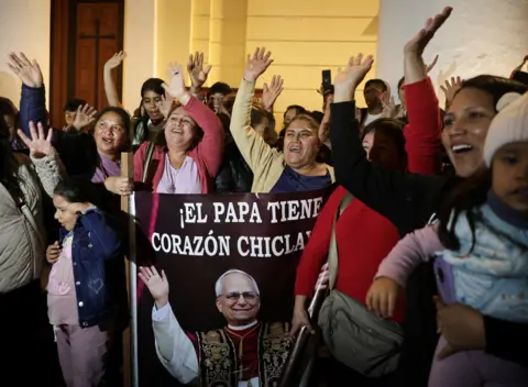 A group of mainly women cheer with their hands in the air and hold a banner of Pope Louis XIV