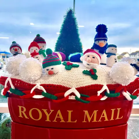 Pat Christie A Royal Mail postbox top with a knitted Christmas top. It has white snowmen and a Santa head. It is stretched across the top of the postbox and the words Royal Mail are in yellow on the red background below.