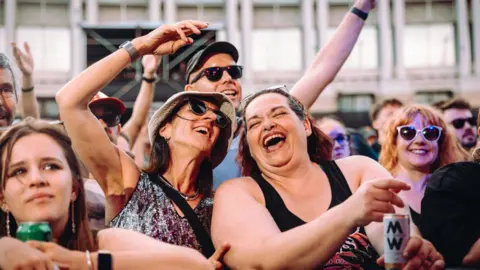 Nadine Ballantyne Two women in a crowd at Bristol Sounds. They are both smiling with their mouths open. One of them has her arm up in the air. Other audience members can be seen by them. Many of them are wearing sunglasses, smiling and holding their arms up.