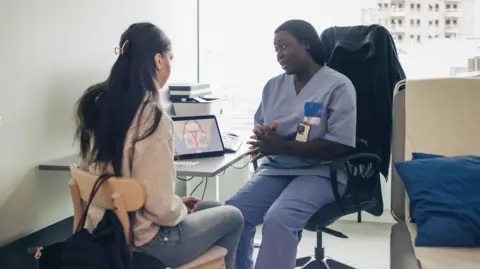 Getty Images A healthcare professional in scrubs explains something to a patient during a consultation, with a diagram of the female reproductive system visible on a laptop screen behind them. The setting appears to be a modern clinical office.