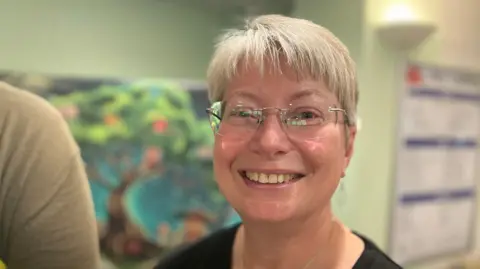 Kate Parish, with short white hair and glasses and wearing a black t-shirt, looking at the camera. A banner showing a tree with books as fruit is blurry but visible in the background.