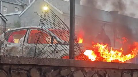 Pacemaker A white car has been set on fire outside a residential area. A hooded figure stands behind the orange flames.
