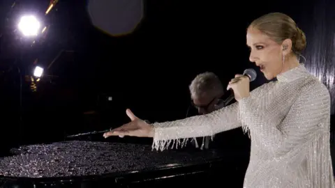 Getty Images Celine Dion performing on stage at the Paris Olympic opening ceremony. She is wearing a long sleeved sparkly silver dress. She's holding a microphone in one hand and the other one is in the air.