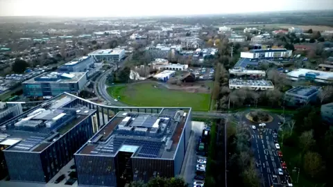 Steve Hubbard/BBC An aerial view of Cambridge Science Park. To the right, cars can be seen entering and leaving the site at a roundabout. There is a large green space in the middle of the picture and it is surrounded by very large industrial buildings and offices. The buildings in the foreground are very modern glass and metal structures but those further back are more dated designs. There are mature trees dotted throughout the site. 