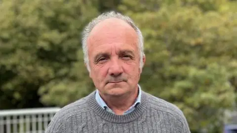 Deputy Neil Inder, a balding man with grey hair looks into the camera. he is wearing a grey jumper with a blue shirt under it. Behind him are green leaves.