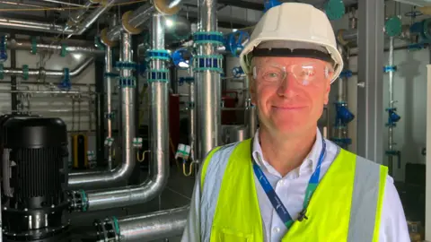 Dave Lees is wearing a hard hat and high-vis jacket and goggles while stood in a room with silver pipes behind him.