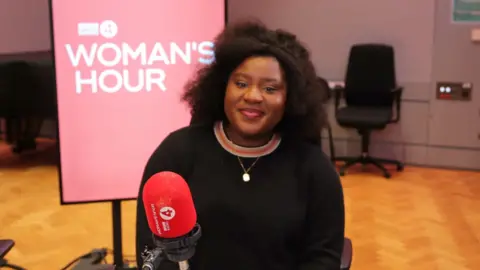 A woman, Susan Wokoma, in the Woman's Hour studio, wearing a black jumper and smiling.