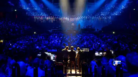 Stuart Nicholl The three band members are on a raised platform in the foreground while the choir line the background. The audience are in between the two and behind the band.