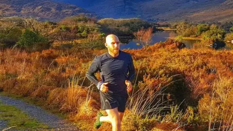Neil Kelders Neil is in a long sleeve black top and black shorts running on a path in front of marshland. He has a bald head and a short beard. In the distance is a river with hills beyond. The light is orange from a low sun at dusk.