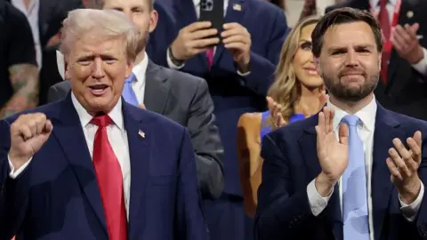 Win McNamee/Getty Images Trump wearing red tie with white plaster bandage on his ear, he is clasping his fist and smiling beside JD Vance in a blue tie clapping and slightly smiling