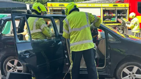 Firefighters cutting two casualties out of a black car. They're in high-viz clothes.