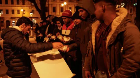 Barbara Zanon/Getty Image A man hands out takeaway food to a line of men in coats and hats, outside in a square in Trieste (February 2024)