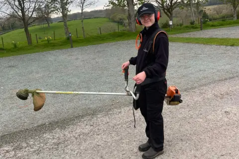 Camping Bible Sarah wearing a black fleece, trousers and work boots. She is standing in front of a field and holding a cordless trimmer. 