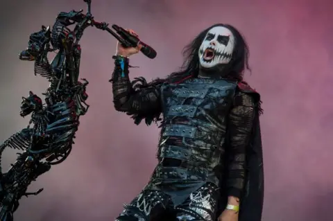 Getty Images A man dressed in black leathers with clown-like face paint. He is singing from a microphone stand made of bones.