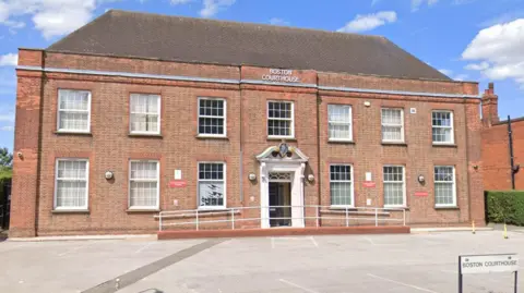 A large two-storey redbrick Georgian-style building. On the front below the roof a signage reads 'BOSTON COURTHOUSE'. Underneath is a window and below that is the front porch which is white and decorative - on the top arch of the door is the court crest logo. There are empty parking bays in front of the building.