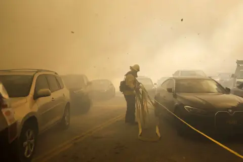 Caroline Brehman/EPA Seorang petugas pemadam kebakaran Los Angeles menyeret selang melewati mobil-mobil yang mencoba melewati jalan berasap di Pacific Palisades.