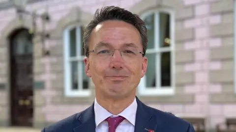 A man with short brown hair and glasses, wearing a navy blue suit, light shift and dark pink tie. He is staring directly at the camera, while standing outside Jersey's States chamber.