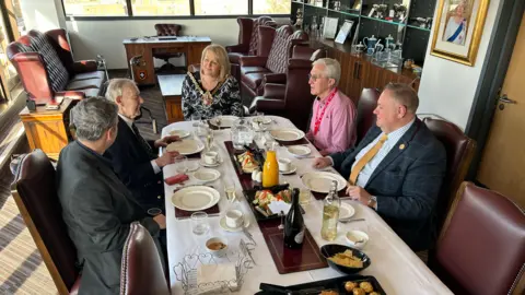 Five people sit around a table with a white tablecloth and food on it. A woman is at the head of the table and there are two men on each side of the table.