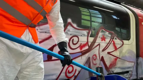 Someone in white coveralls and hi viz cleaning graffiti from a train