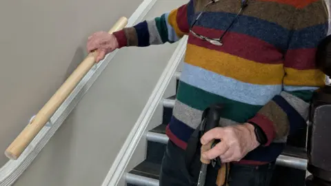 Julie Mariotti/BBC A close-up picture of a man standing at the end of a staircase holding up a handrail prototype against a wall to demonstrate how it would look if it were installed. He is holding onto his stick with the other hand.