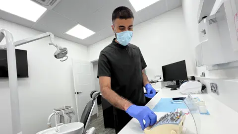 A man with black hair wearing a grey dental tunic wears blue protective gloves and a facemask as he carries out a task in a dental surgery. The room has white walls and ceiling, a dentist chair and small sink, and white counters.