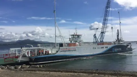 A small car ferry docks on a lough shore on a sunny day in 2017. The boat is painted white, navy and blue with a Carlingfordferry.com sign on the site. A green ramp allows vehicles to roll on and off the boat. The outline of the Mourne Mountains can be seen in the background. 