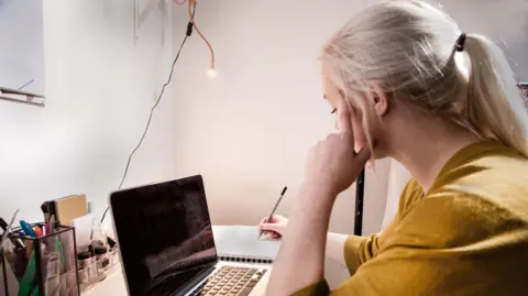A student sits at home at a desk, in front of a laptop. She is holding a pen and writing on paper. She has blonde hair in a ponytail and is wearing a mustard jumper. There are pens on her desk in a pen pot.