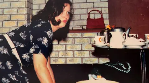 Sheringham Theatre Ruth Jones, when she was 26, with dark wavy hair, red lipstick and in a black and white floral dress. She is on her knees cleaning and looking away from the camera. There is a pile of crockery and teapots on a nearby table and a red handbag on a shelf against a fake brick wall.
