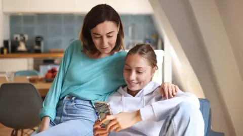A mum and daughter sit together on a blue sofa in a bright room, looking at a smartphone. The mum, who has dark brown hair and wears a turquoise top with blue jeans, has her arm around her daughter's shoulder. The daughter has fair hair tied back and wears a grey hoodie and holds the phone. A kitchen with white cupboards, blue wall tiles, wooden worktops, chairs, appliances and a fruit bowl is blurred in the background.