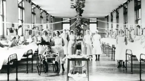 Alistair Ritch A black-and-white image of a hospital ward dressed for Christmas, with streamers stretching out from a central foliage display. Children sit up in rows of beds against each opposing wall, wearing party hats. The nurses, wearing white pinafores and hats, stand by the beds, posing for the photo. A young girl in a wheelchair is at the front of the picture.