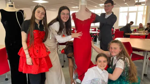 BBC (Left to right) Secondary pupils Deyka, Dennisa and Jude (front row) Zayda and Myla from Brook Mead Academy, who attended a De Montfort University upcycling workshop, wearing glamorous prom outfits, standing next to mannequins with dresses on and looking at the camera.