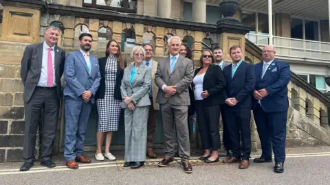 Group of people stood smiling infront of stone steps to Matlock County Hall building