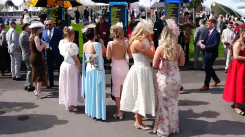 PA Media A group of women in formal dresses stand with their back to the camera studying the odds at Aintree