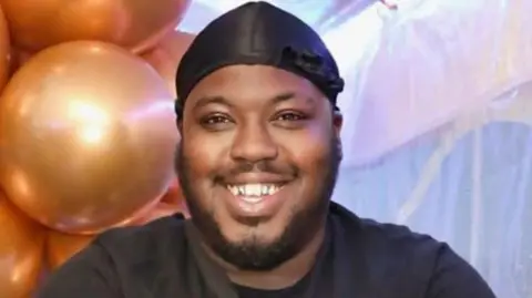 Family A man with a black bandana and black shirt, smiles. He is in front of some orange balloons and some light blue and pink decorations.