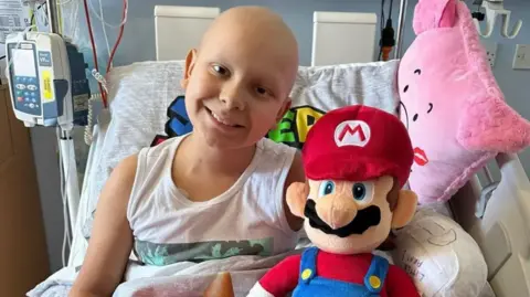 Family handout Rian in a hospital bed with medical machines around him, smiling and holding a Mario cuddly toy