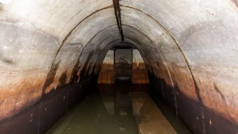 The inside of a WW2 air raid shelter with a bench on the right submerged in water that covers the floor. There are orange and brown marks up the walls of the shelter which has an arched shape.