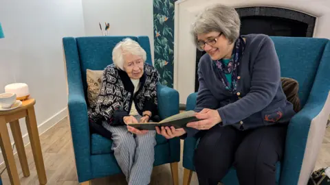 Shirley Bartlett, on the left, wearing a cardigan and blue and white striped trousers. She is sat on a blue armchair. To her right is Ruth Brighton wearing a blue cardigan and navy blue trousers. She is also sat in a blue armchair. There are both holding a photo album book between them and looking at it. 