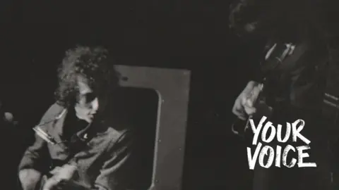 Bob Dylan with his distinctive curly hair, has his harmonica round his neck and is playing his electric guitar at Manchester Free Trade Hall