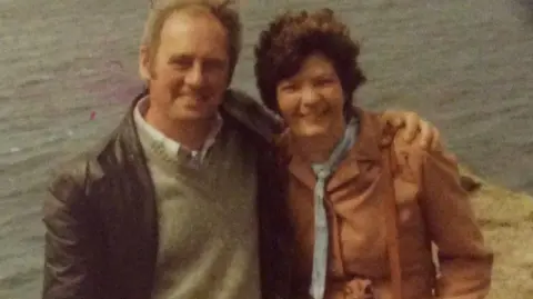 Don Bullock A faded older photo of a couple. The man has a 70s style brown leather jacket and grey jumper. His hair is receding. He has his arm around a woman with short dark hair and wearing an orange mac. The sea is behind them.