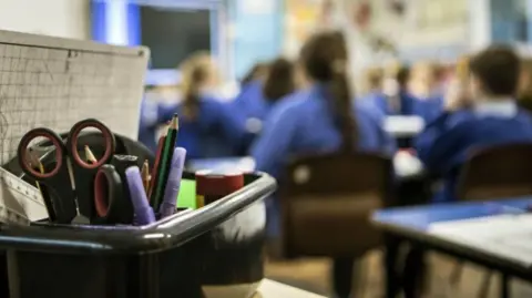 A box of stationery - including scissors and pencils - with primary school pupils in the background.
