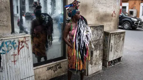 AFP via Getty Images A female vendor stands on a street corner in Luanda. She is wearing a headscarf and vest-top, plus an African-print skirt. She has assorted colourful shoe laces over her shoulder.