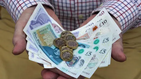 Man holding pound coins and notes in five, ten, twenty and fifty denominations, in both hands.