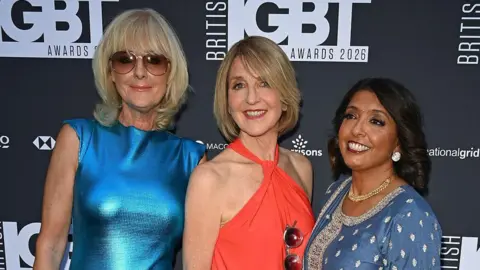 Getty Images A photograph of Jane Moore, Kaye Adams and Sunetra Sarker from the waist up on the red carpet. The background is black with a logo reading 'British LGBT Awards'. Jane, on the left, wears a shiny blue dress and sunglasses with her hair in a blond bob. Kaye wears a red halter-neck dress with a similar hairstyle. Sunetra is on the right and wears a blue dress with gems, a gold necklace and silver earrings. She has dark hair.